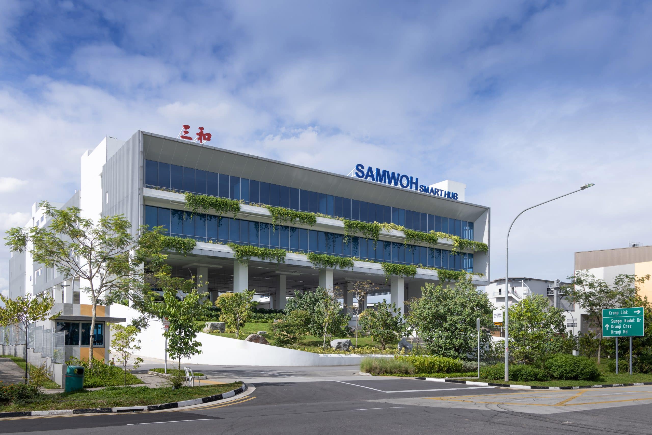 xterior view of the Samwoh Smart Hub in Singapore, showcasing its modern architecture. The first positive energy industrial building features solar panels on the roof, lush greenery surrounding the structure, and large windows that reflect natural light, emphasising its eco-friendly design.