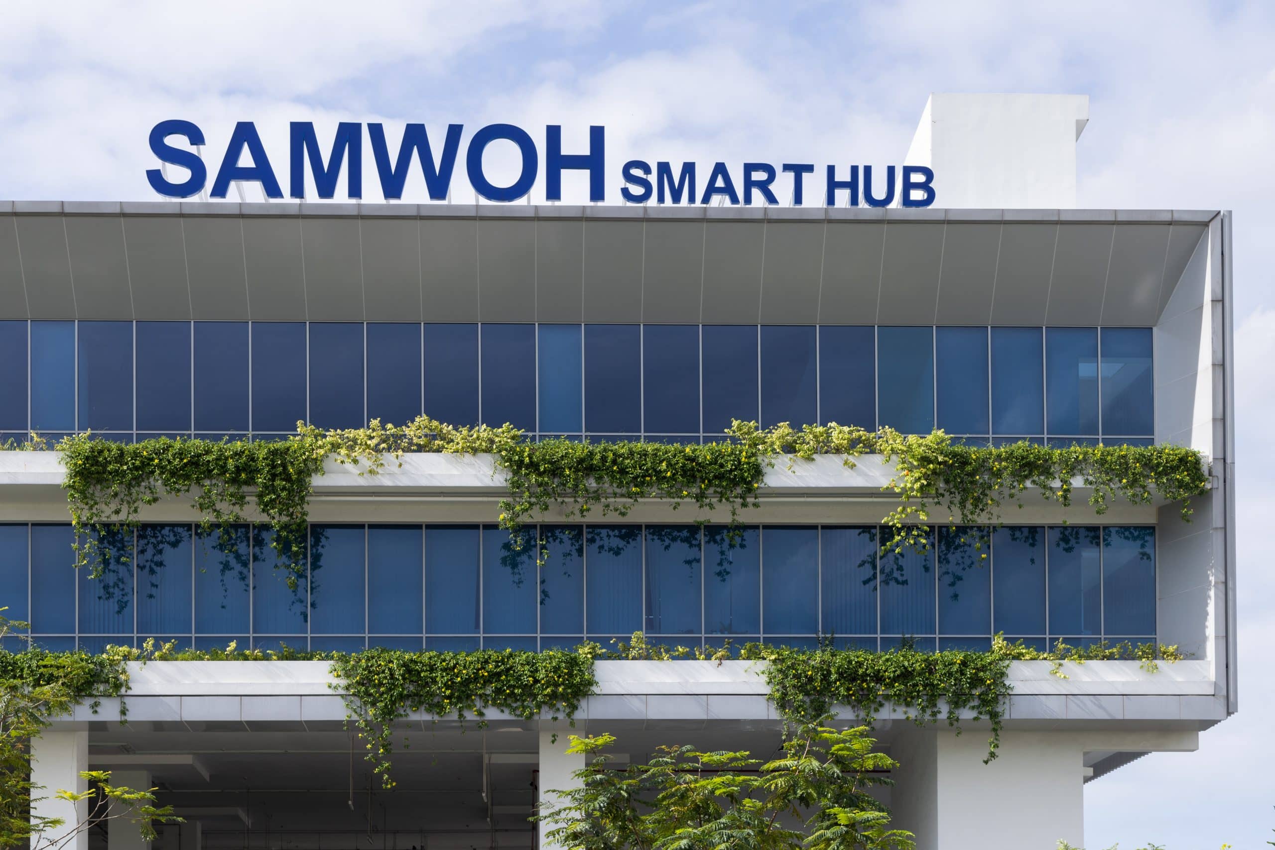 Exterior view of the Samwoh Smart Hub in Singapore, showcasing its modern architecture. The first positive energy industrial building features solar panels on the roof, lush greenery surrounding the structure, and large windows that reflect natural light, emphasising its eco-friendly design.