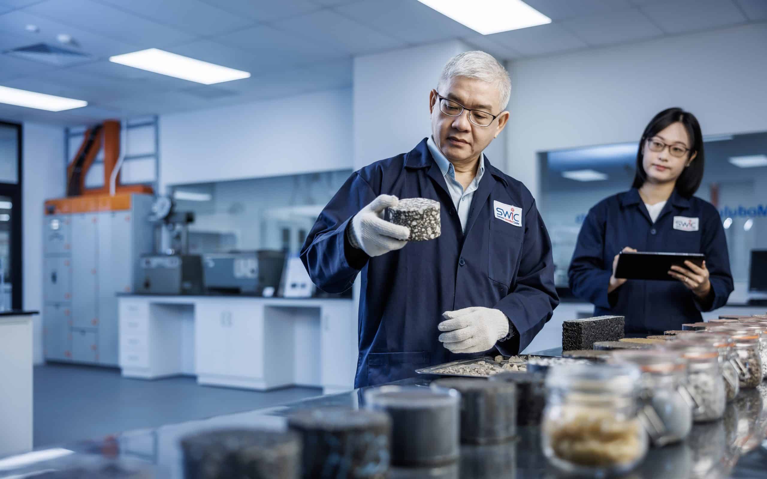 Two lab researchers examining an asphalt sample, one holding the sample up for closer inspection while the other takes notes. They are dressed in lab coats, surrounded by testing equipment and research materials in a well-lit laboratory setting.