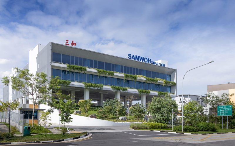xterior view of the Samwoh Smart Hub in Singapore, showcasing its modern architecture. The first positive energy industrial building features solar panels on the roof, lush greenery surrounding the structure, and large windows that reflect natural light, emphasising its eco-friendly design.